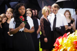 A crowd gathers to mourn at a funeral. A woman in front clutches roses to her chest.