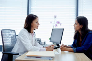 Doctor sits at desk listening to person with long dark hair in distress on other side of desk