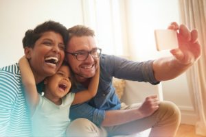 Family with young child laughs as they take a selfie with a smartphone.