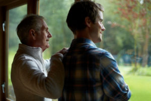 Thoughtful parent and adult son stand at window of cottage and look out toward woods