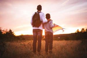 Rear view photo of father with arm around son's shoulders at sunset