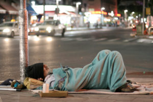 Night photo of a homeless man lying on the sidewalk under a teal blanket.