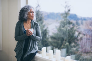 Person with curly, shoulder-length gray hair holds cup of tea and looks out large window toward trees, smiling slightly