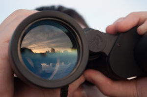 A reflection of the sky appears in a pair of binoculars held by an unseen man.