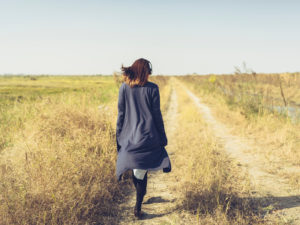 Rear view photo of person with long hair wearing long sweater and headphones walking along path in yellow grassy field 