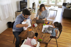 Family sits at the table; the mother is on her laptop and the father is reading to the children.