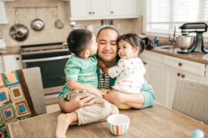 Parent sits at table and hugs two happy toddlers sitting on table in tidy kitchen