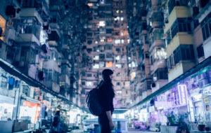 Person with backpack and dark hair stands downtown looking up at skyscrapers