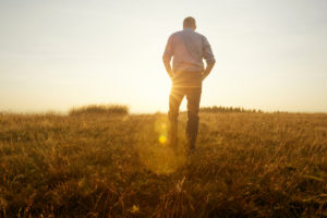 Rear view of person with short hair in pants and sweater walking in field at sunset