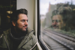 Adult with short hair and facial hair looks out window thoughtfully, smiling slightly