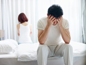 A couple, both who have faces down or turned away, sit on opposite ends of bed