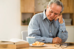 A sad man sits alone in his kitchen with a croissant and a cup of tea.