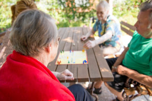 A senior woman plays a board game with three other people on a picnic table.