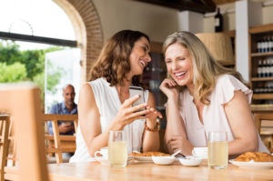 Two cheerful mature adults with long hair smiling and laughing at something on phone at table over breakfast in cafe