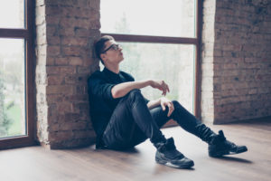 Young adult with short hair and glasses sits on floor against wall, legs out, looking up thoughtfully with serious expression