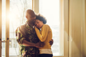Sunlight shines through the window, illuminating a soldier and wife embracing.