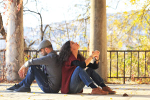 A young couple sits back to back. There are some stone columns and trees in the background.