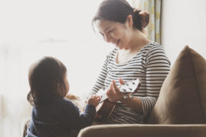 Mother and toddler sit on sofa. Mother plays guitar and holds it for child.