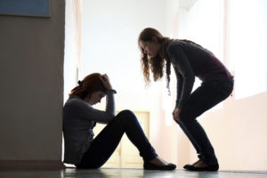 Person sits on the floor looking down, while friend leans over to offer support