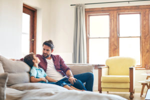 Parent with facial hair sits on sofa with child having a serious talk but smiling