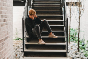 Young adult sits on stairs outside house with hands covering face looking down