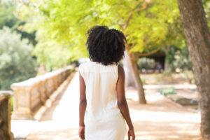 Rear view of person of color in white dress with natural hair walking along street under trees