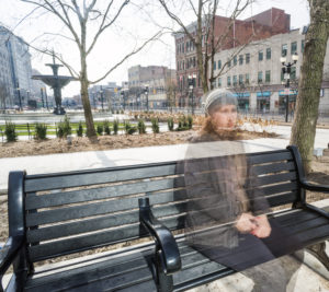 Half-transparent photo of person with curly hair wearing hat and coat sitting on bench on cold day