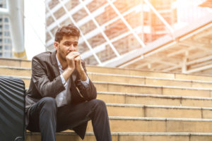 Young adult with briefcase and short hair sits on outdoor steps outside building, hands folded under chin, looking thoughtful but distressed