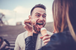 A bearded man makes a silly face as his friend feeds him ice cream.