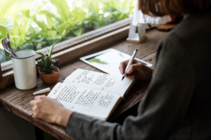 A woman writes in her journal while sitting at a desk surrounded by greenery.