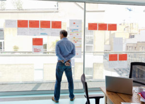 Older adult in business shirt and pants looks at glass wall covered with notes, hands behind back