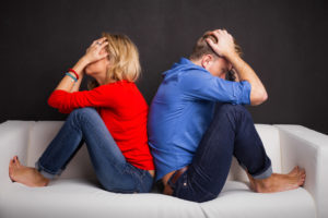 Adult with long blonde hair in red shirt and jeans sits with back to other adult in jeans and blue shirt. Both have face in hands and irritated demeanor