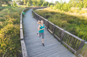 Adult with blonde hair in ponytail runs along bridge path in tree-filled park