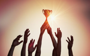 Silhouette of a trophy held above a crowd of reaching hands.