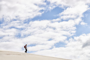 Man walking down pale sand hill with expansive cloudy sky in the background