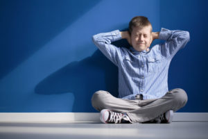 A young boy in a blue shirt sits against a blue wall, covering his ears with both hands.