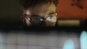 A man looks at a computer screen, which reflects in his glasses