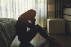 Woman sits on the floor at the foot of a bed with her head in her hands.