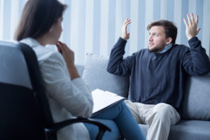 A therapist sits in the foreground, listening to a man discuss his frustrations.