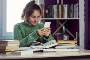 Donna seduta alla scrivania, procrastinare con uno smartphone accanto ai libri di testo aperti