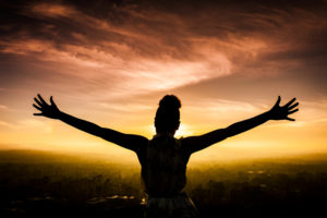 Woman faces sunset and raises arms in triumph.
