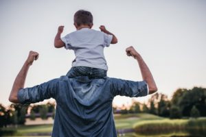 Toddler son sitting on his dad's shoulders, mimicking his arm motions
