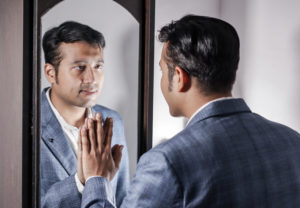 A man in a suit stares at his reflection while touching the mirror.