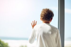 Boy in white shirt looks out of a window.