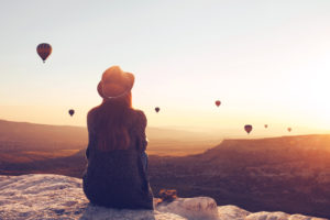 A young woman sits and watches hot air balloons drifting across the horizon.