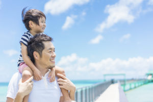 A young boy rides his father's shoulders as they walk down a sunny pier.