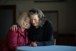 Two senior women hug while sitting at a small library table.