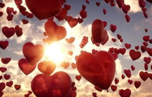 Red heart balloons floating in sky with sun in background.