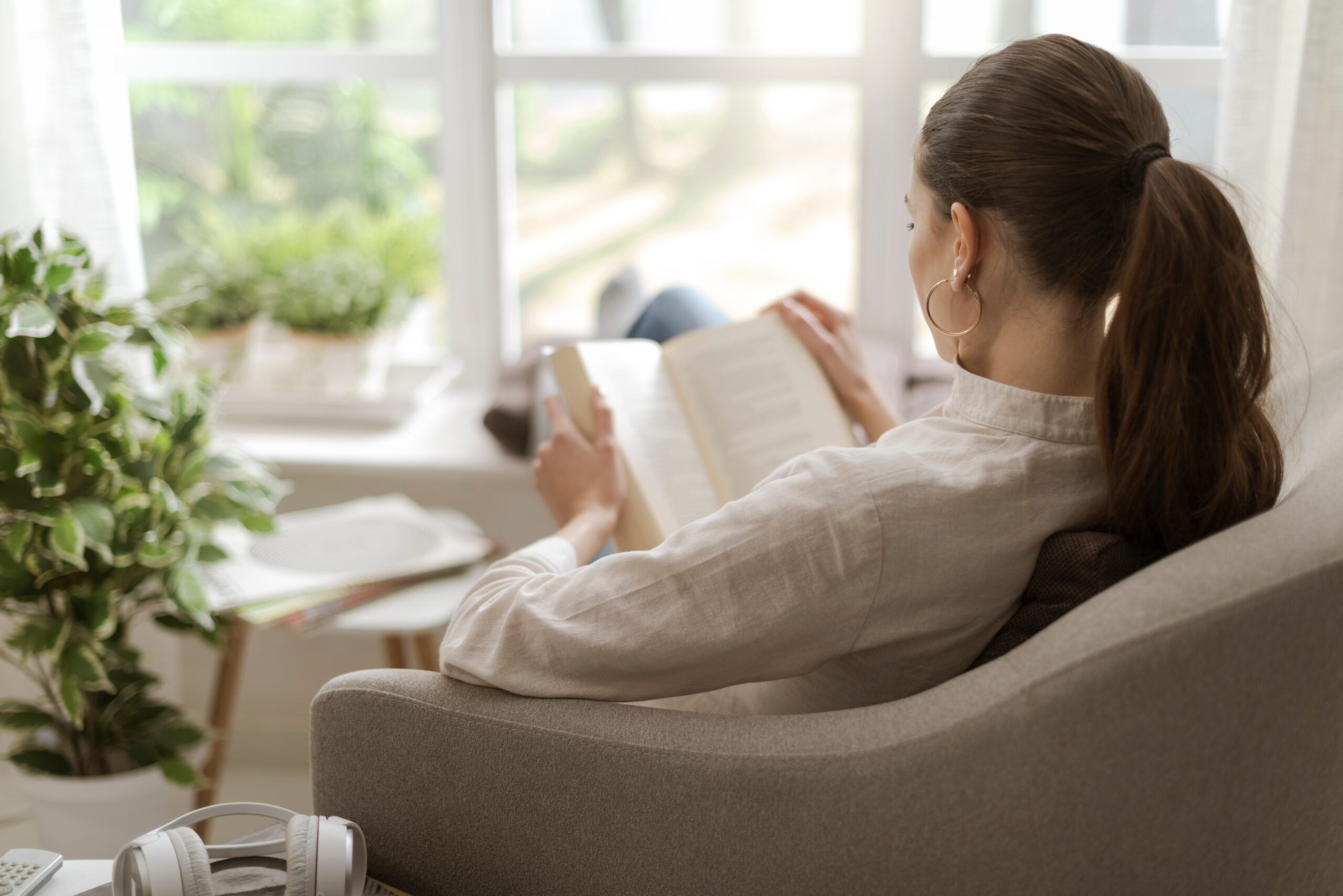 Woman relaxing at home next to a window, sitting in an armchair and reading a book
