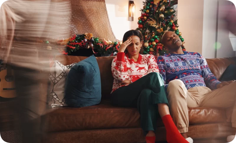 Understanding Winter Depression and Seasonal Affective Disorder: It’s Not Just the Weather 1 A stressed woman with a hand on her forehead and a sleeping man on a couch, surrounded by festive holiday decor, illustrating understanding winter depression.